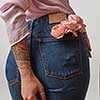 Beautiful female wearing in jeans with fresh roses of living coral color in a back pocket and pink shirt on a light gray background. Place for text. Concept of Woman's or Mother's Day.