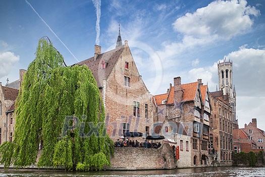 Bruges, Belgium - May 17, 2012: Water channel with old houses, a bell tower in the background and a cafe on a spring day against the blue sky. Bruges, Belgium - May 17, 2012: Water channel with old houses, a bell tower in the background and a cafe on a spring day against the blue sky.