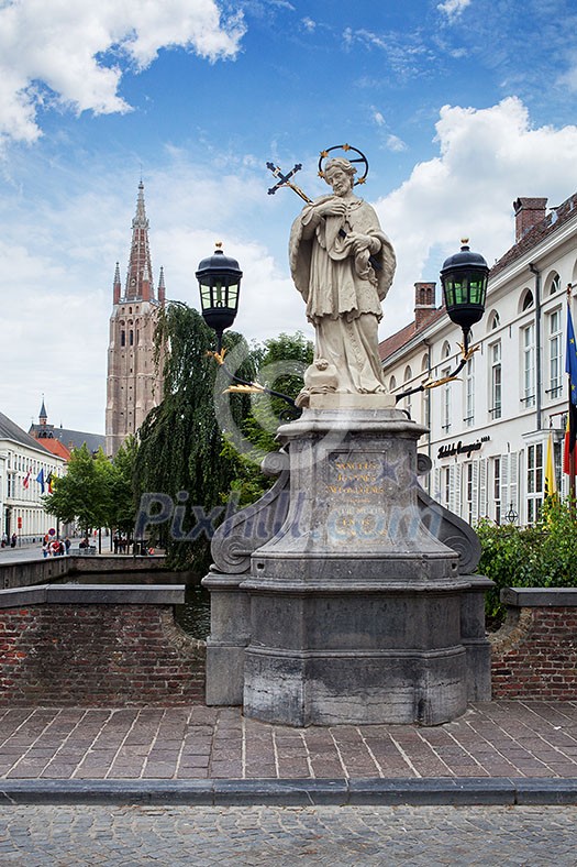 Bruges, Belgium - August 16, 2013: Scenic cityscape with statue of Johannes Nepomucenus, church tower against the blue sky in Bruges