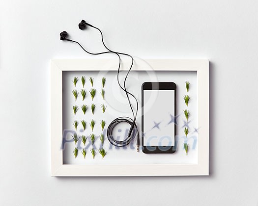Creative ogranic herbal pattern of pine needles twigs, mobile smartphone with blank screen for mockup and headphones in a frame on a light gray background. Place for text. Flat lay.