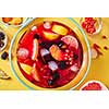 Drink with different berries and fruits with ice cubes in a glass jug presented on a yellow background. Top view