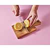 On a wooden board the hands of a woman cut a ripe lemon for making a vitamin drink around a pink background with space for text.
