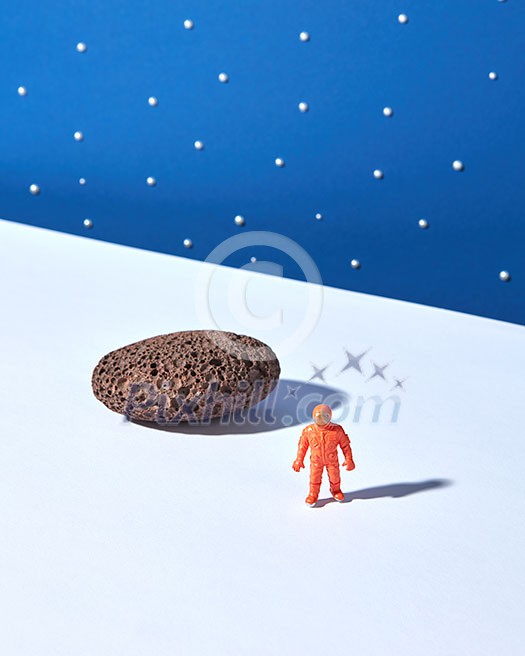 Toy plastic astronaut on a light ground with stone and shadows against blue starry sky background. Place for text. Outer space concept. Toy plastic astronaut on a light ground with stone and shadows against blue starry sky background. Place for text. Outer space concept.