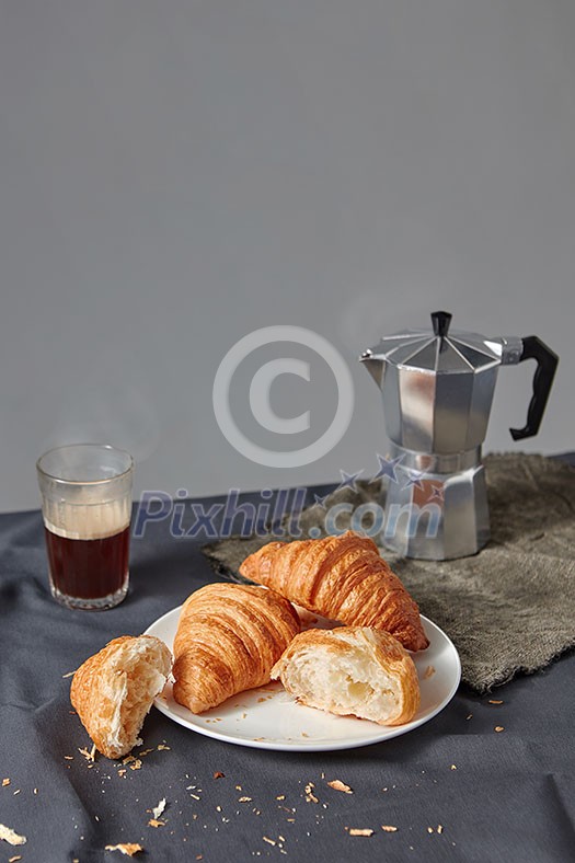 Glass of aromatic hot coffee and plate with freshly baked french croissants on a grey background. Continental breakfast concept. Place for text. Glass of aromatic hot coffee and plate with freshly baked french croissants on a grey background. Continental breakfast concept. Place for text.