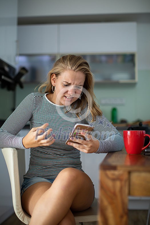 Teenage girl using her cell phone, being angry, while having a cup of tea in modern kitchen setting (shalllow DOF, color toned image)