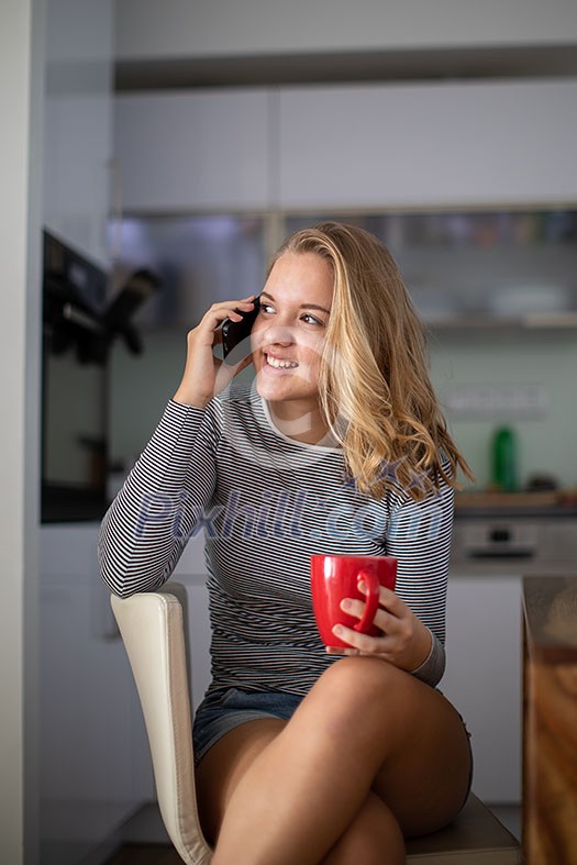 Teenage girl calling on her cell phone while having a cup of tea in modern kitchen setting (shalllow DOF, color toned image)