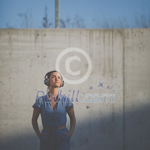 Pretty, young woman with headphones standing against concrete wall, enjoyig the hi-fi quality sound, listening to her favorite songs (color toned image) Pretty, young woman with headphones standing against concrete wall, enjoyig the hi-fi quality sound, listening to her favorite songs (color toned image)