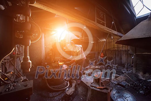 traditional blacksmith manually forging the red hot molten metal on the anvil with sunlight through the windows in smithy workshop. Blacksmith working metal with hammer in the forge traditional blacksmith manually forging the red hot molten metal on the anvil with sunlight through the windows in smithy workshop. Blacksmith working metal with hammer in the forge