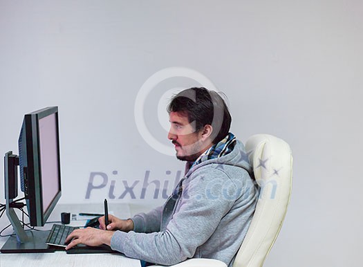Young Graphic Designer Working at Workplace. Guy Sitting at Table Retouching Photo Using Digital Tablet with Pen and desktop computer Indoors. Freelancing and Distance Job Concept Young Graphic Designer Working at Workplace. Guy Sitting at Table Retouching Photo Using Digital Tablet with Pen and desktop computer Indoors. Freelancing and Distance Job Concept