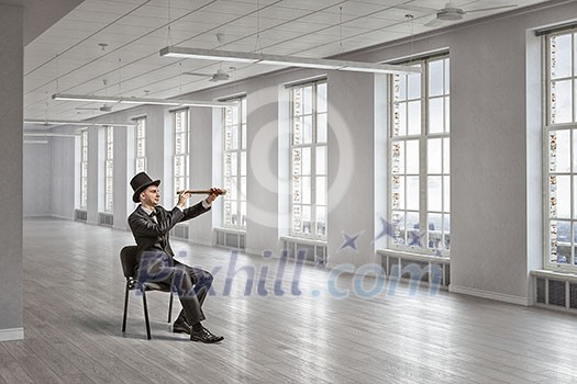 Young businessman wearing cylinder in chair looking in spyglass