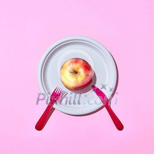 Natural organic apple on a disposable plate served with plastic knife and fork on a pink background with copy space. Top view.
