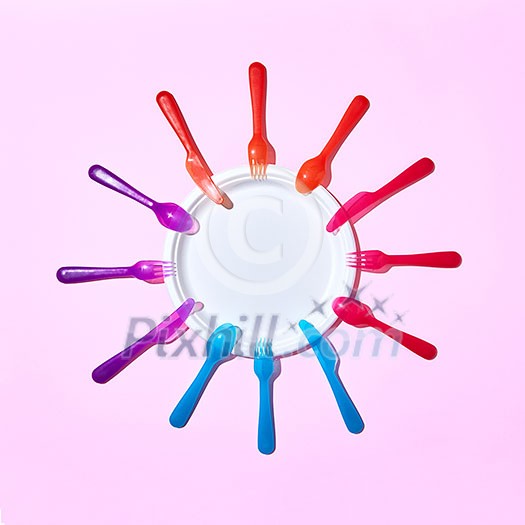 Round frame from colorful disposable plastic forks and knives around white paper empry plate on a pastel pink background with copy space. Round frame from colorful disposable plastic forks and knives around white paper empry plate on a pastel pink background with copy space.