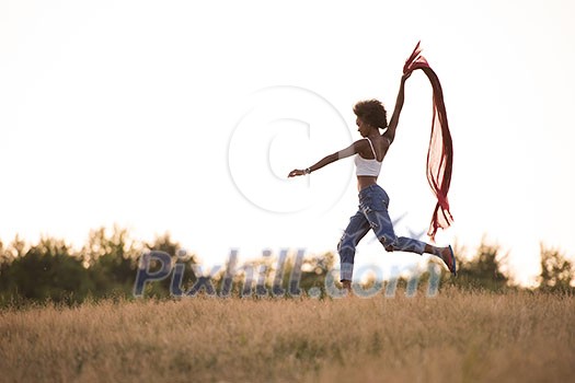 Young beautiful black girl laughs and dances outdoors with a scarf in her hands in a meadow during sunset