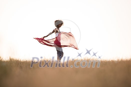 Young beautiful black girl laughs and dances outdoors with a scarf in her hands in a meadow during sunset