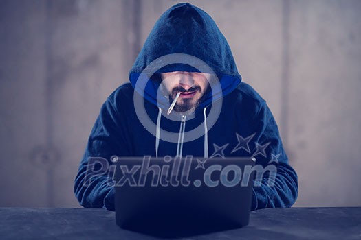 Young talented hacker smoking cigarette while working on laptop computer in dark office with concrete wall in the background