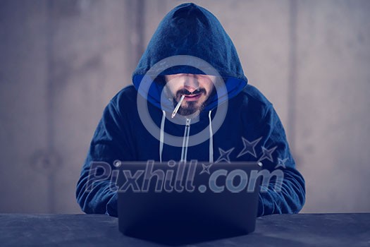 Young talented hacker smoking cigarette while working on laptop computer in dark office with concrete wall in the background Young talented hacker smoking cigarette while working on laptop computer in dark office with concrete wall in the background