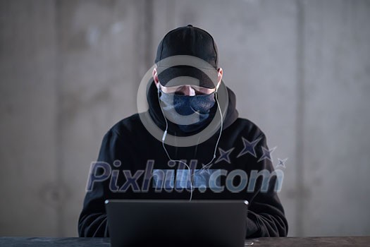 Young talented hacker using laptop computer while working in dark office with concrete wall in the background