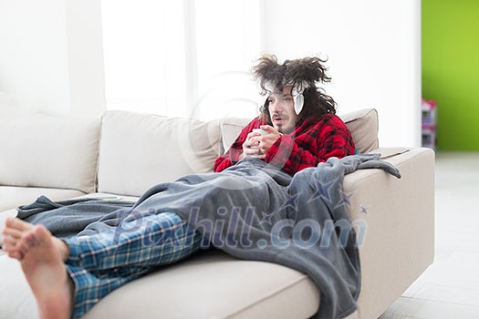 young Man in bathrobe with flu and fever wrapped holding cup of healing tea while sitting on sofa at home