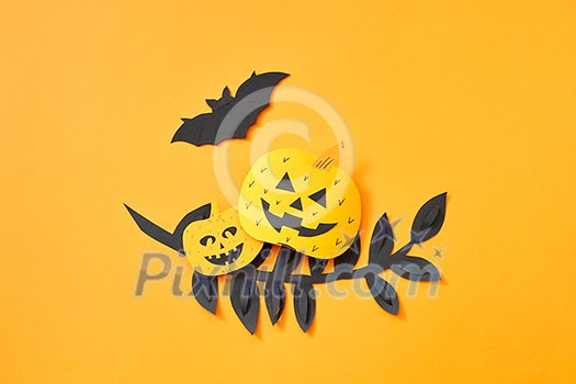 Black paper leaves branch, flying bat and pumpkin with a scary face on an orange background with space for text. Handcraft paper composition to Halloween. Flat lay