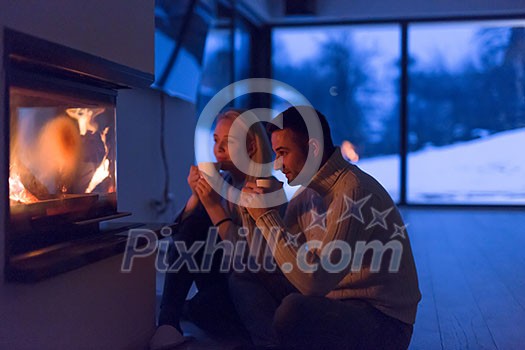 Young romantic couple sitting on the floor in front of fireplace,talking and drinking tea on cold winter night at home