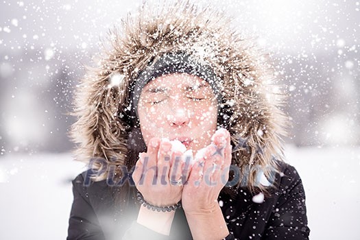 Happy young woman wearing winter clothes while blowing snow on snowy day with snowflakes around her in beautiful winter forest