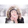 Happy young woman wearing winter clothes while blowing snow on snowy day with snowflakes around her in beautiful winter forest