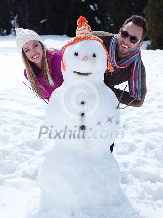 portrait of happy young couple with snowman at beautiful winter day with fresh snow and forest in background