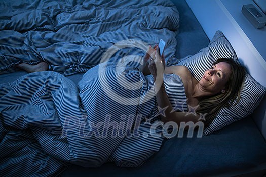 Pretty, middle-aged woman using her phone before sleep in bed in the evening