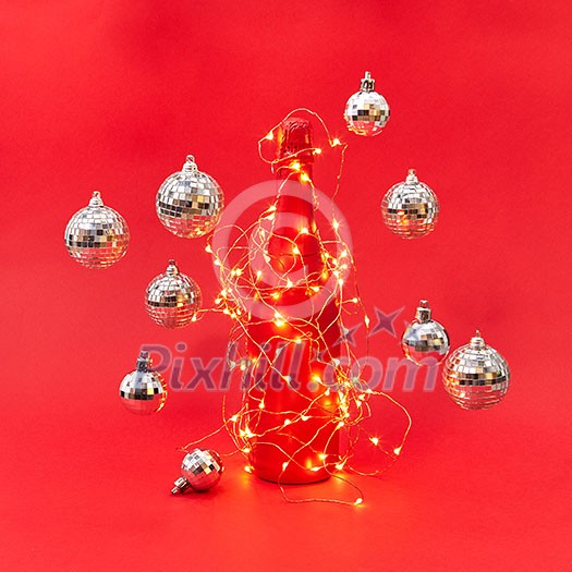 New Year shined lights string on a creative painted wine bottle and floating balls around on a red background with copy space. Christmas congratulation card.
