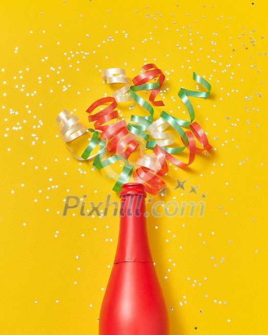 Holiday red painted wine bottle with multicolored paper srirals as a champagne bubbles on an yellow background, copy space. Flat lay.