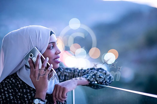 Young modern Muslim woman wearing scarf veil on urban city  street at night texting on smartphone with bokeh city light in background