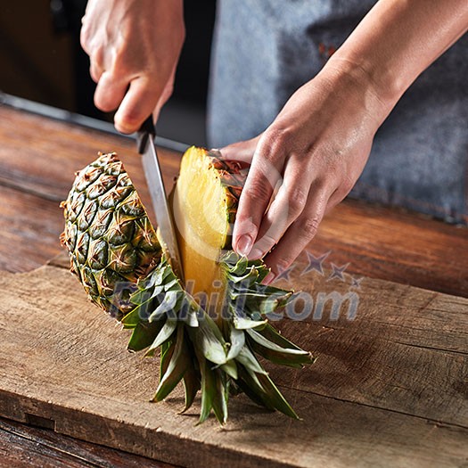 Woman's hands with a knife cut a juicy pineapple on a wooden board on a brown table around a dark background. Healthy ingredient