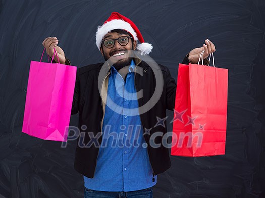 Indian  man wearing party clothes in Santa hat with shopping bag on dark  background studio dark skinned middle eastern Santa Claus merry Christmas
