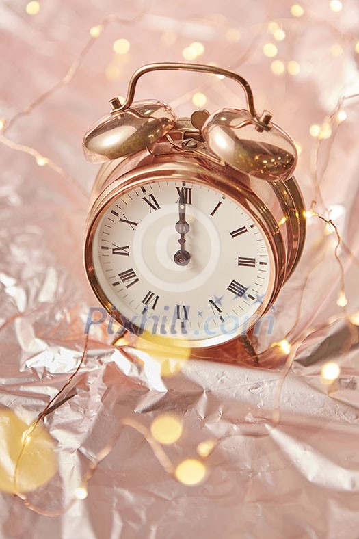 Close up golden painted alarm clock with Christmas time is midnight on a shiny golden abstract background with lights garland, copy space.