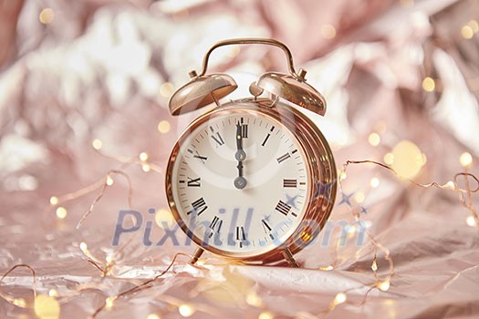 Golden alarmclock with time is midnight on a shiny golden abstract background with string of lights. Greeting card.