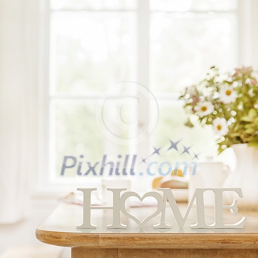 Home 3D text standing on wooden kitchen table top with blurred background showing breakfast tablewear, windowframe and a vase filled with garden flowers. Space for text.