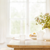 Home wooden kitchen table top with focus in front and blurred background showing breakfast tablewear, windowframe and a vase filled with garden flowers. Space for text.