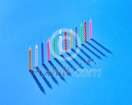 Greeting birthday card from vertically standing candles for sweet dessert and cakes on blue background with hard shadows, one of them is burning, copy space. Greeting birthday card from vertically standing candles for sweet dessert and cakes on blue background with hard shadows, one of them is burning, copy space.
