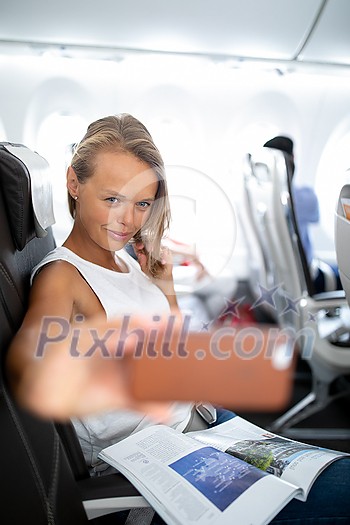 Young happy woman taking selfie photo with passport document sitting on the aircraft seat near a window during the flight in the airplane