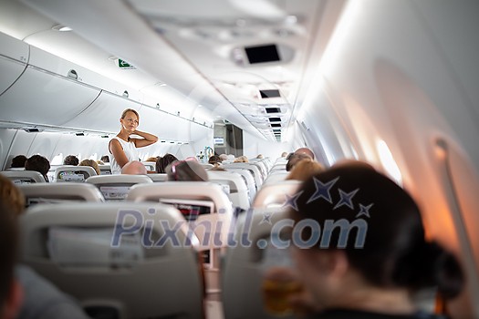 Pretty, young happy woman aboard an airplane during a lang haul commercial flight - stretching her legs and sore neck a bit, walking in the aisle