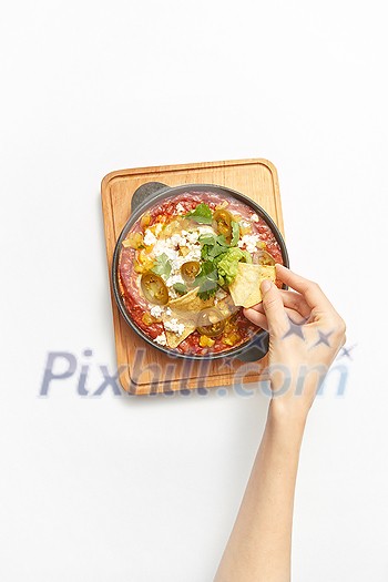 Freshly cooked traditional dish Shakshuka from fried eggs with tomatoes, pepper, vegetables and herbs in a pan with woman's hand above wooden board on a light grey background, copy space. Top view.