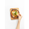 Freshly cooked traditional dish Shakshuka from fried eggs with tomatoes, pepper, vegetables and herbs in a pan with woman's hand above wooden board on a light grey background, copy space. Top view.