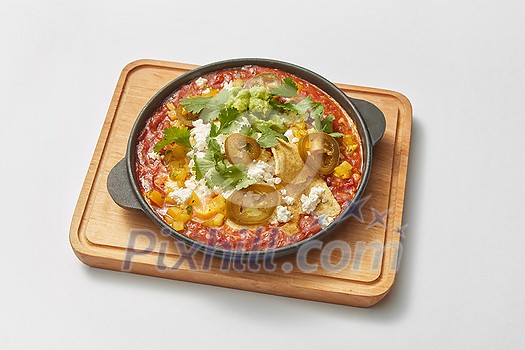 Homemade shakshouka dish cooked from fresh fried eggs with tomatoes, pepper, vegetables and herbs in a pan on a wooden board on a light grey background, copy space. Middle eastern traditional dish.