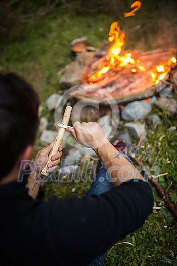 Young man making tools out of wood by a campfire Young man making tools out of wood by a campfire