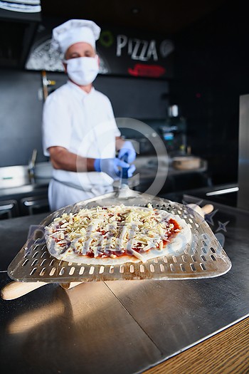 Skilled chef preparing traditional italian pizza in interior of modern restaurant kitchen with special wood-fired oven. Wearing protective medical face mask and gloves in coronavirus new normal concept Skilled chef preparing traditional italian pizza in interior of modern restaurant kitchen with special wood-fired oven. Wearing protective medical face mask and gloves in coronavirus new normal concept
