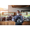 waiter in a medical protective mask serves  the coffee in restaurant durin coronavirus pandemic representing new normal concept