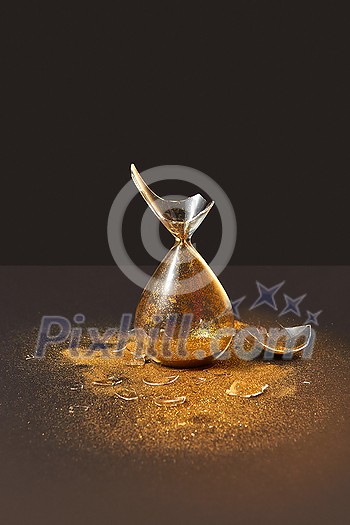 Old-fashioned vintage vertically standing crashed sandglass with bits of glass and golden sand on a duotone dark background, copy space. Old-fashioned vintage vertically standing crashed sandglass with bits of glass and golden sand on a duotone dark background, copy space.