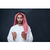 young arabian man in traditional clothes making traditional prayer to God, keeps hands in praying gesture in front of black chalkboard representing modern islam fashion and ramadan kareem concept