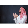 young arabian man in traditional clothes making traditional prayer to God, keeps hands in praying gesture in front of black chalkboard representing modern islam fashion and ramadan kareem concept