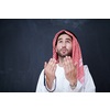 young arabian man in traditional clothes making traditional prayer to God, keeps hands in praying gesture in front of black chalkboard representing modern islam fashion and ramadan kareem concept